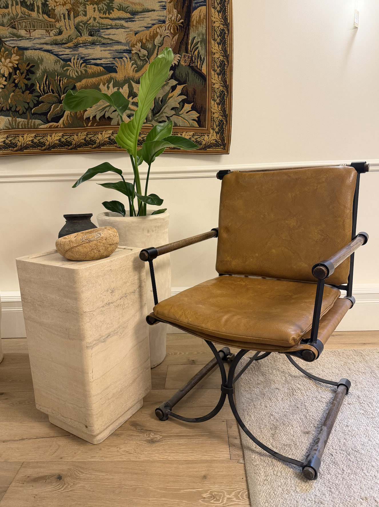 A brown leather and metal framed lounge chair next to a beige stone pedestal with decorative objects and a potted plant, with a tapestry hanging on the wall behind.