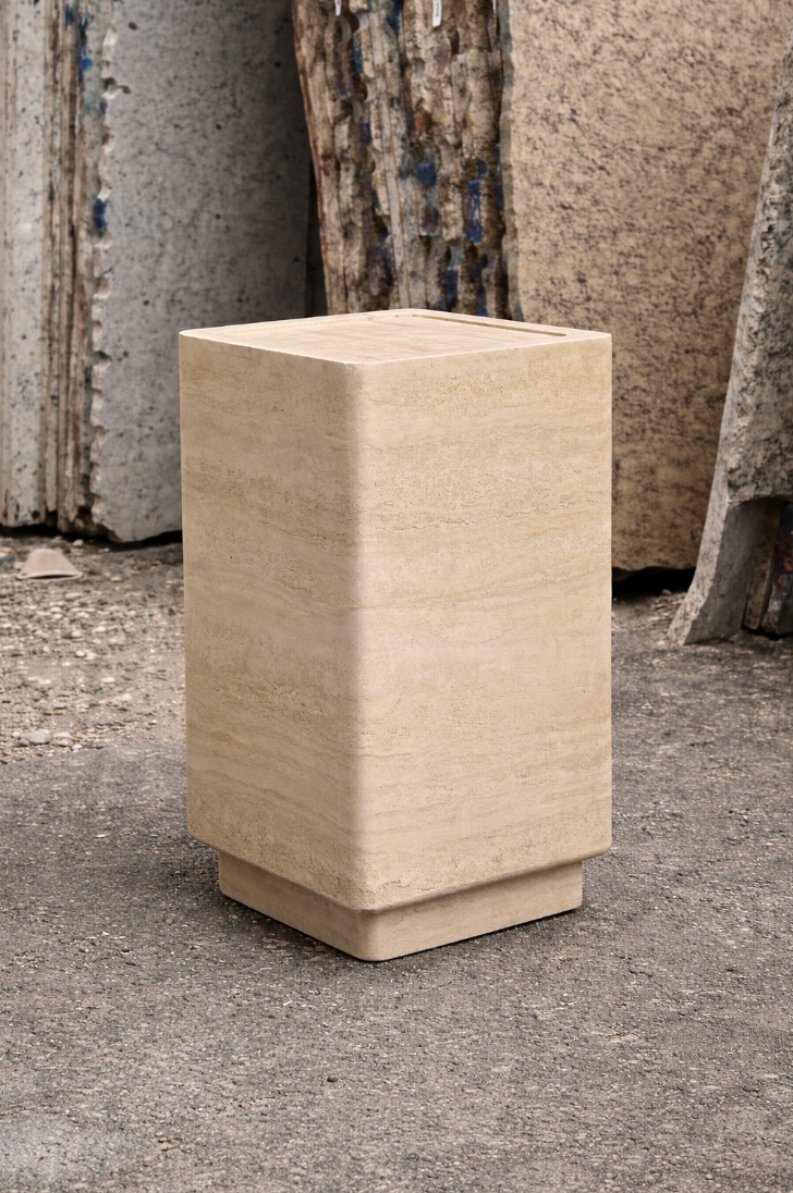 A beige stone cube with rounded edges stands on a concrete surface, with stacked stone slabs in the background.
