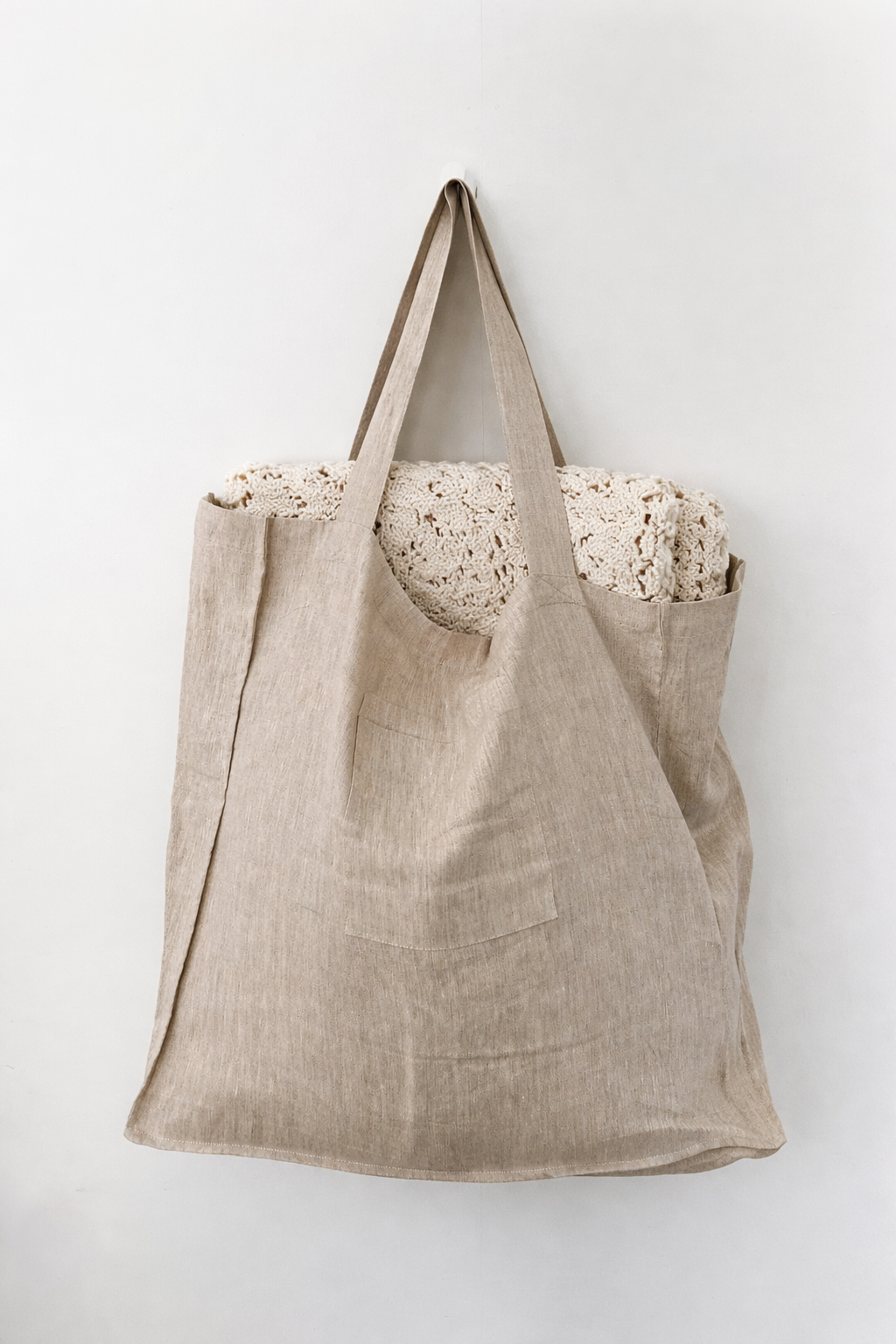 Beige fabric tote bag hanging on a wall hook with a chunky knit blanket inside.