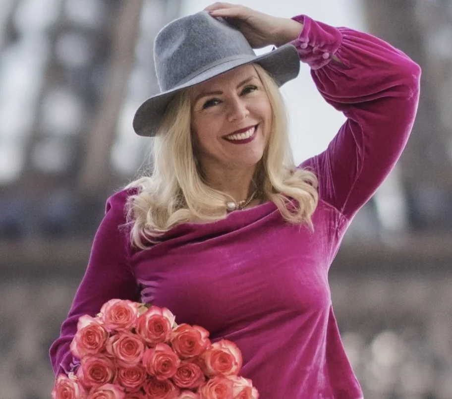 A woman with blonde hair, wearing a pink velvet top and a gray wide-brimmed hat, smiling and holding a bouquet of pink roses outdoors.