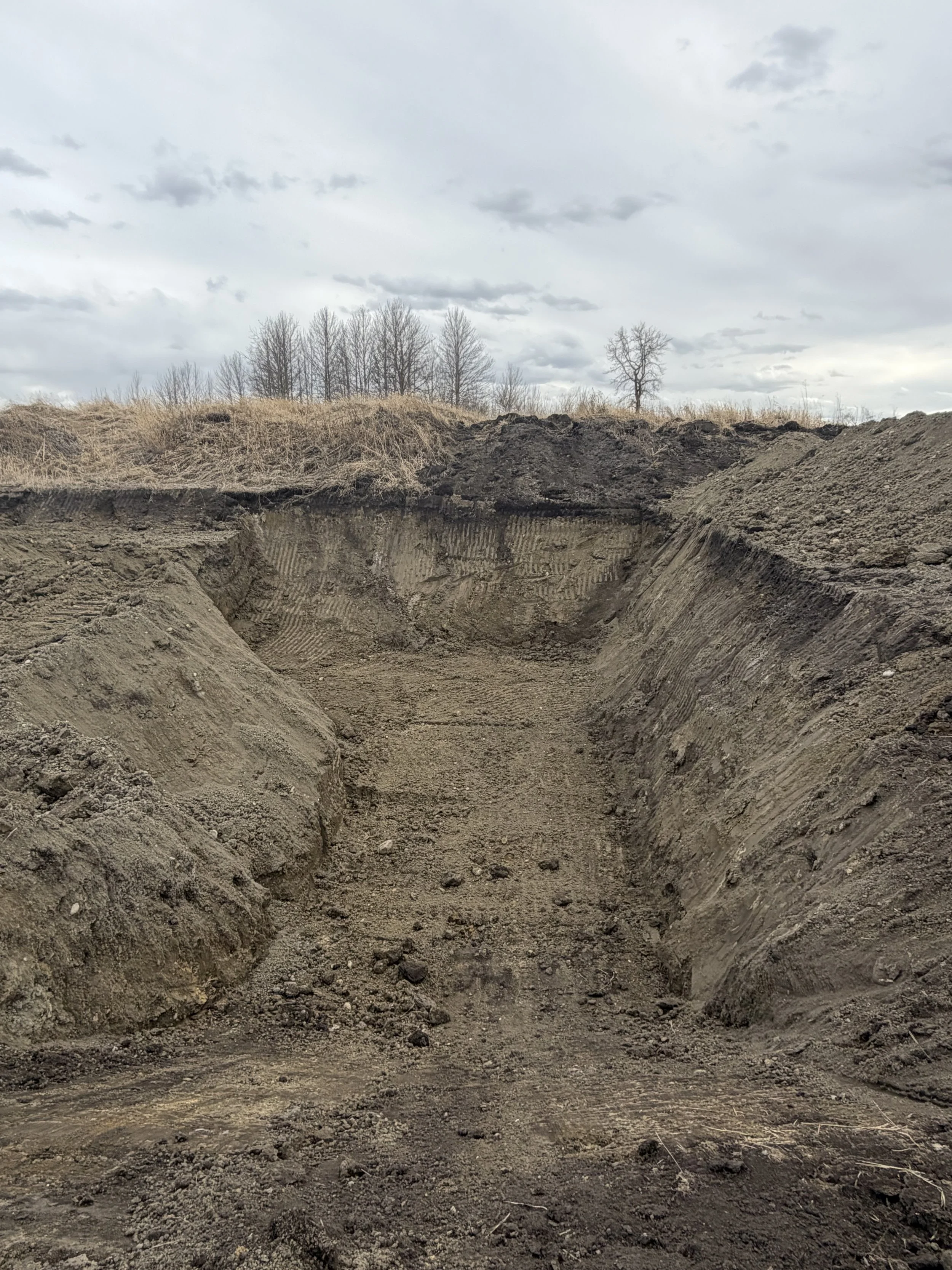 A large excavation site with deep dirt trenches, barren soil, and a cloudy sky in the background.