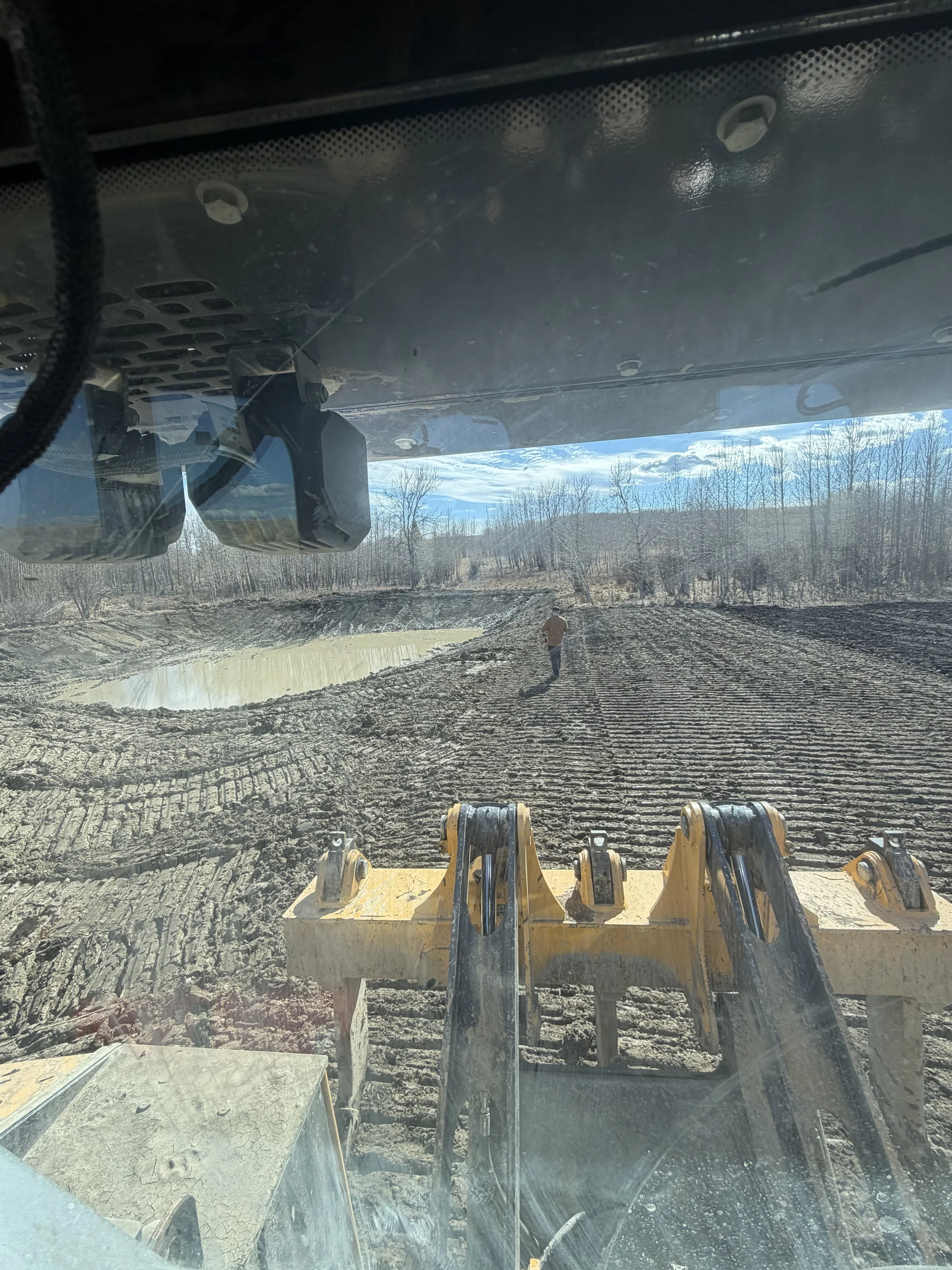 View from inside a construction vehicle cab showing a person walking on uneven dirt terrain with tire tracks, a water puddle, trees, and a partly cloudy sky in the distance.