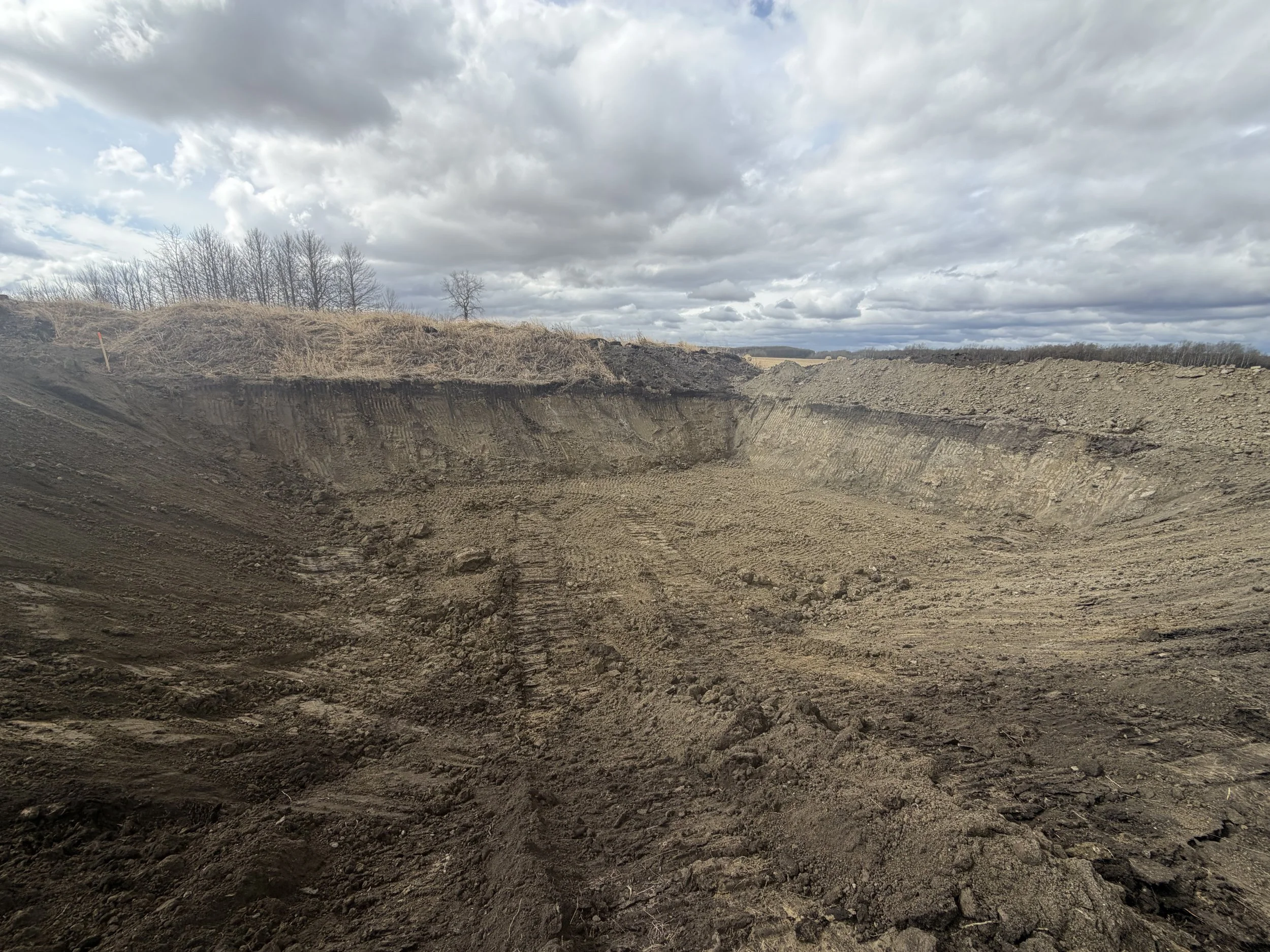 A large excavation site or crater with exposed dirt layers, under a cloudy sky with some trees on the horizon.