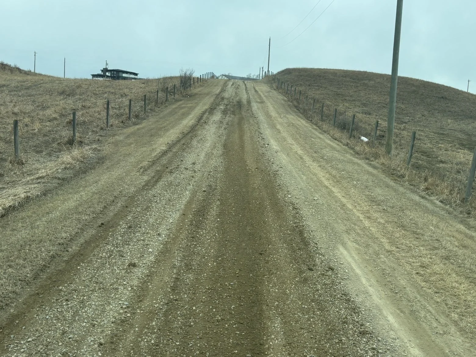 Dirt gravel road leading uphill between fenced sides, with a house and utility poles in the distance under cloudy sky.