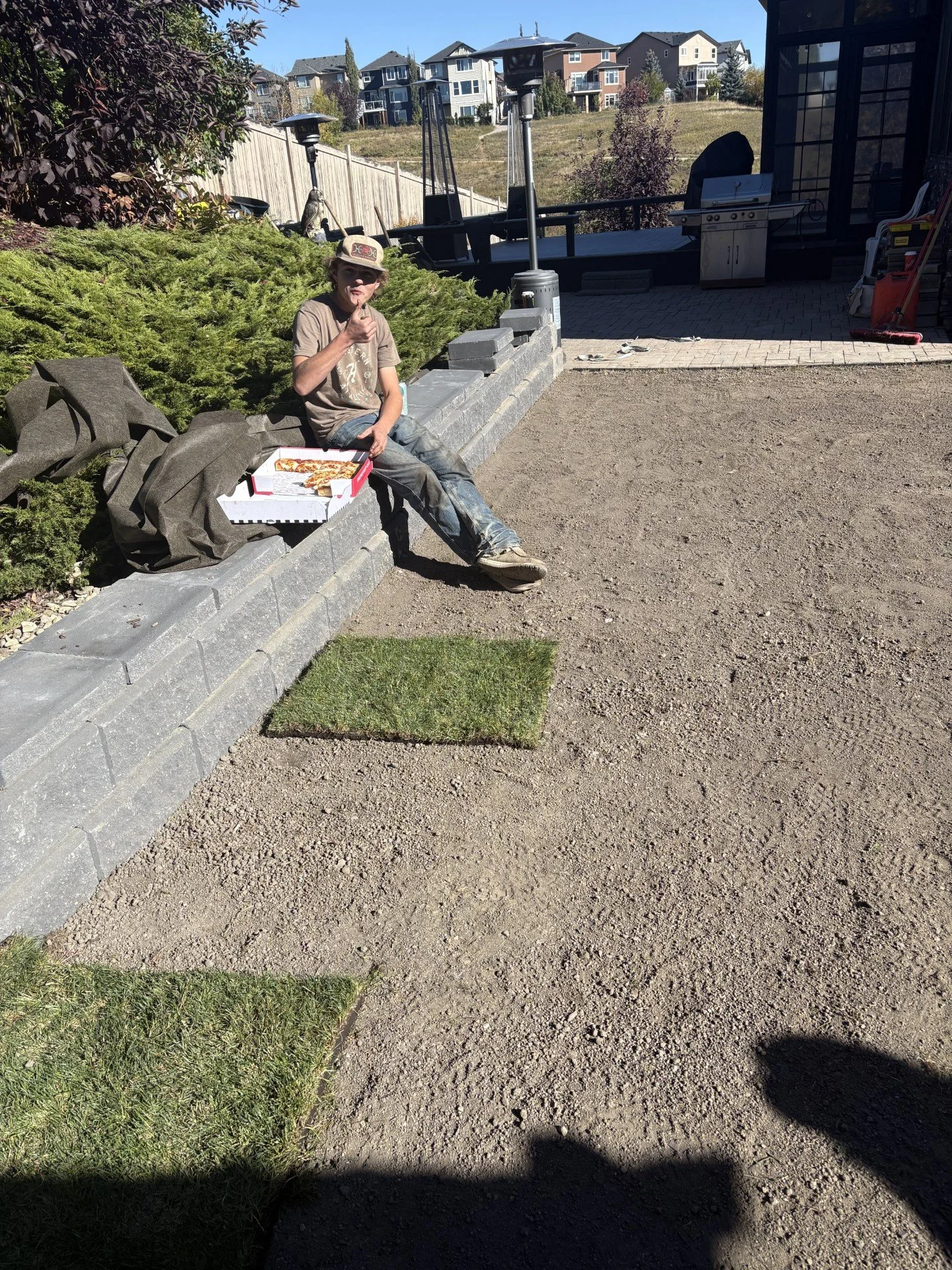 Child sitting on a low stone wall eating pizza outdoors in a backyard with a flagstone patio, grill, and houses in the background.