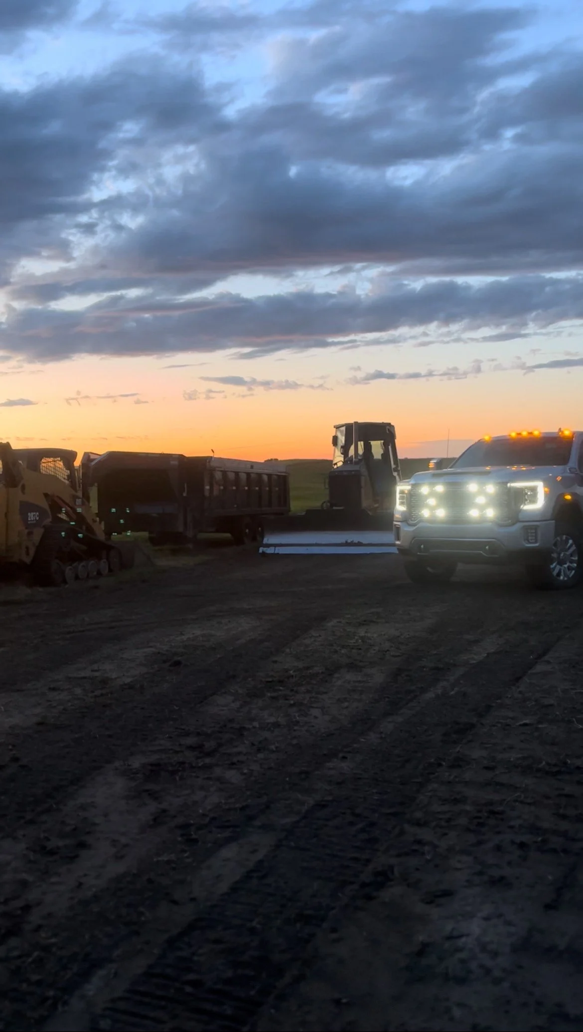 Construction vehicles parked on a dirt lot during sunset, with a cloudy sky overhead.