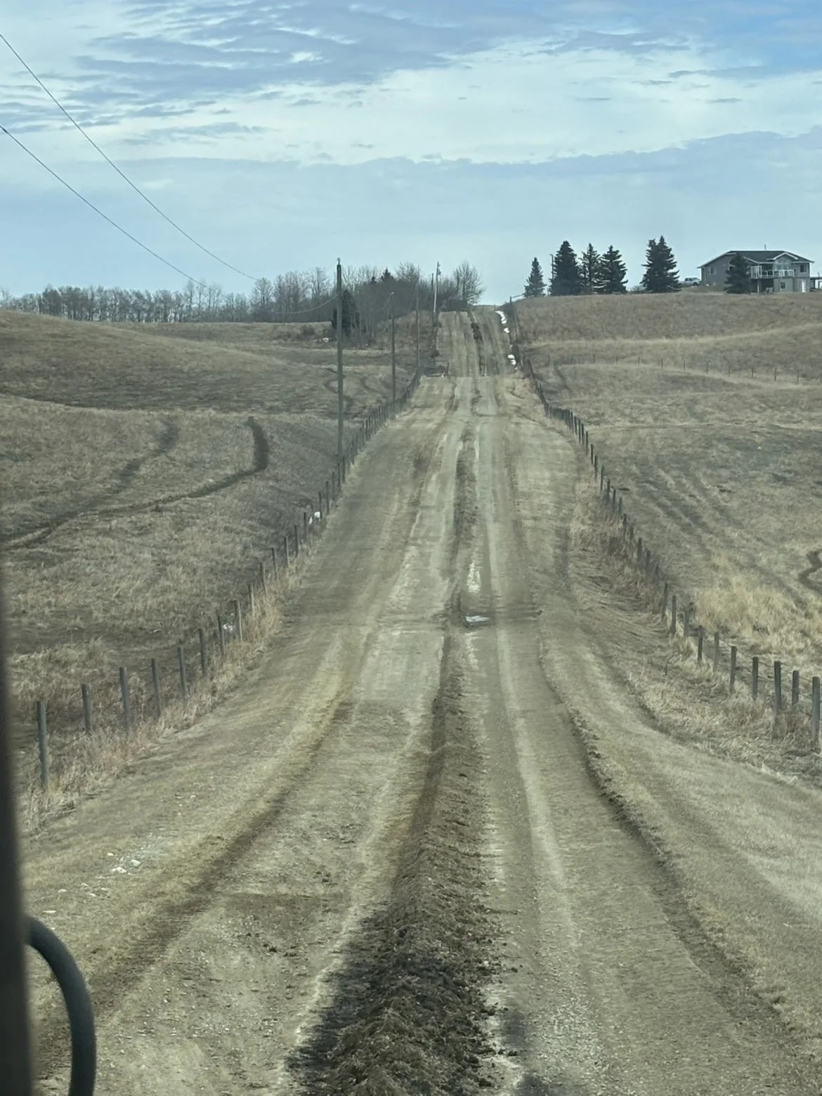 Dirt country road stretching into the distance, flanked by fences and farmland, under a partly cloudy sky with some trees and houses on the horizon.