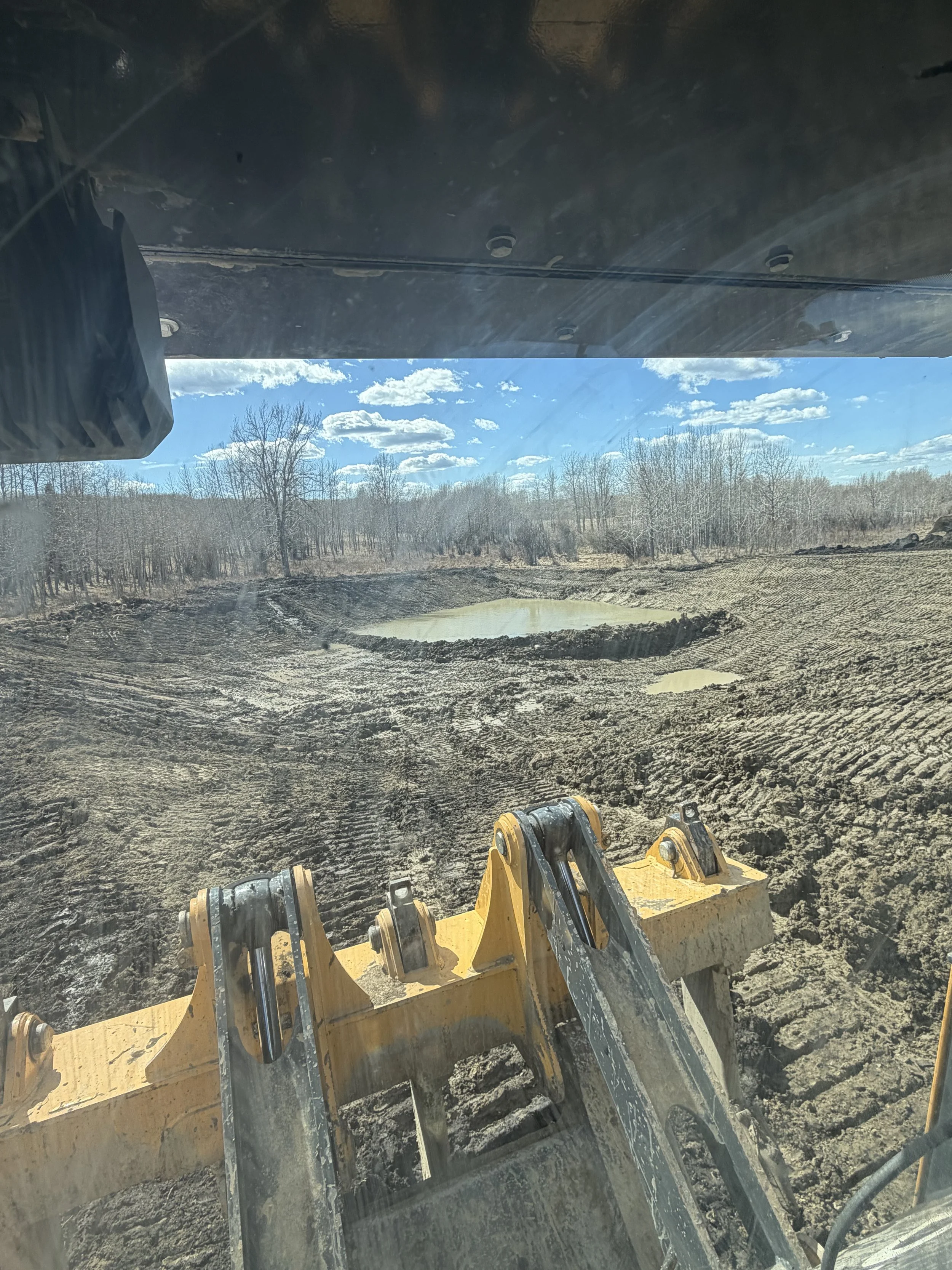 View from inside a construction vehicle looking out over a muddy construction site with a small pond, trees, and blue sky with clouds in the background.