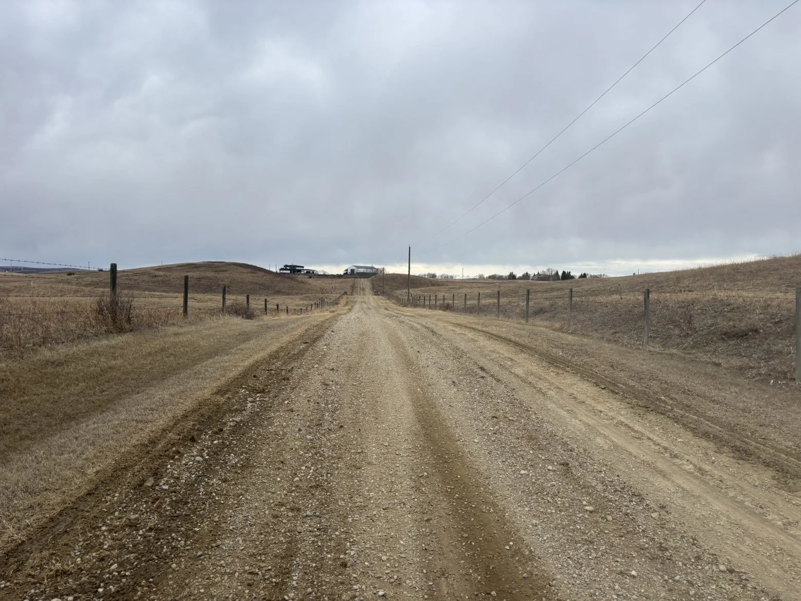 A gravel country road lined with wooden fence posts on both sides, leading to a distant farmstead under a gray, overcast sky.