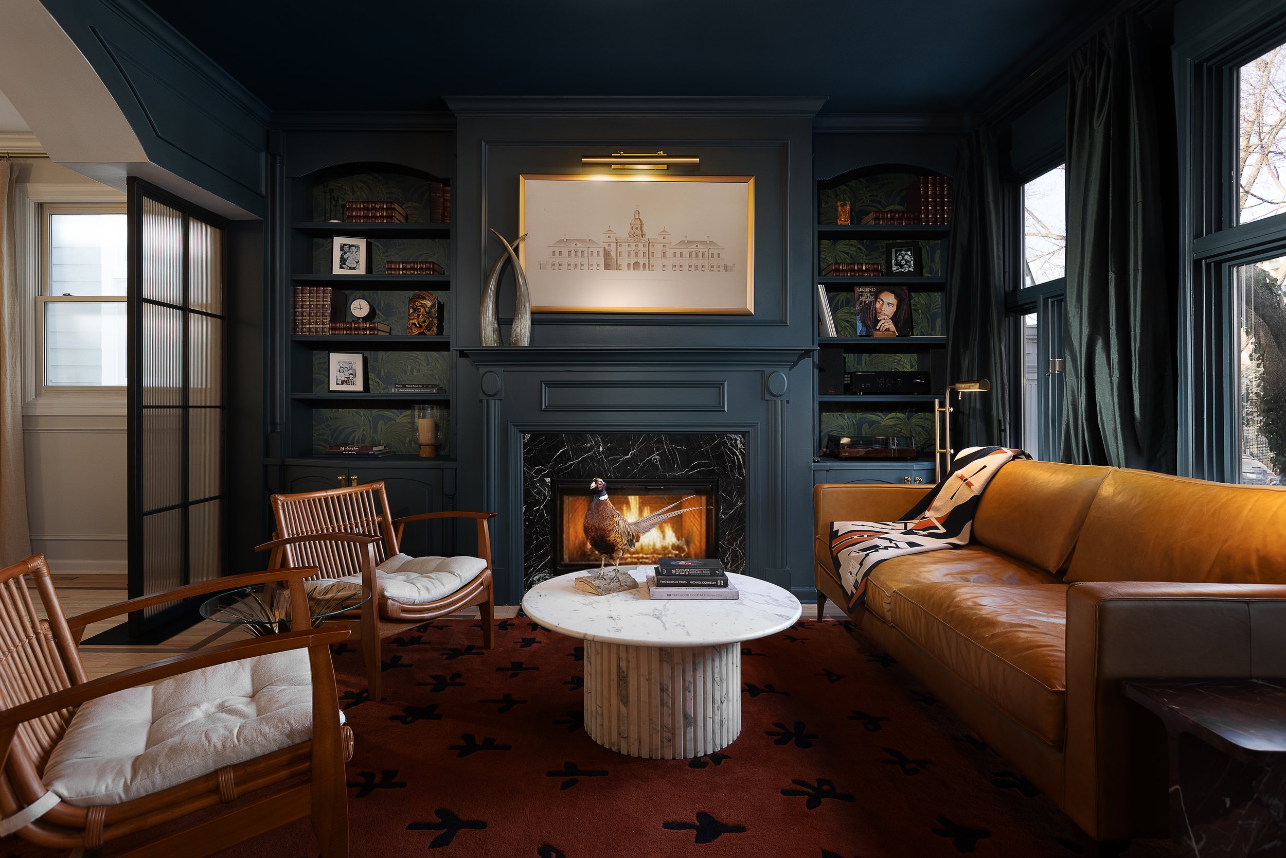A cozy living room with dark blue walls, a fireplace with a black marble mantel, and built-in bookshelves. There is a mustard-colored leather sofa, two wooden armchairs with white cushions, and a round marble coffee table with a decorative turkey figurine and stacked books. Large windows with dark curtains allow natural light to fill the space.