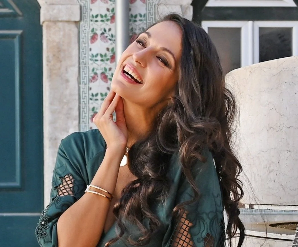 A woman with long, dark, curly hair smiling and touching her face with her hand, outdoors in front of a portuguese tiled wall. 