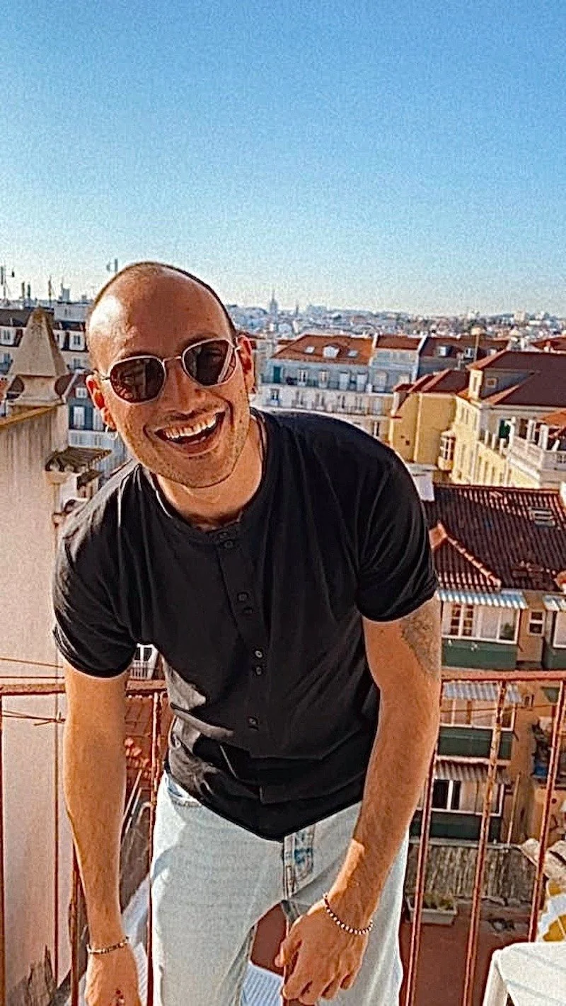A smiling man wearing sunglasses, a black shirt, and light-colored jeans leaning on a balcony with a cityscape of rooftops and a clear blue sky in the background.