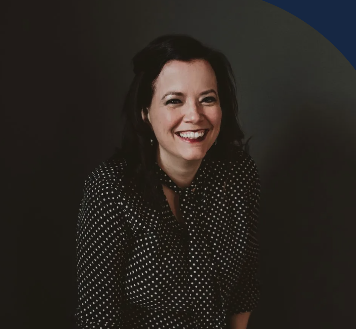 A smiling woman with short light brown hair and a goatee, wearing a black collared shirt, against a blue-green gradient background.