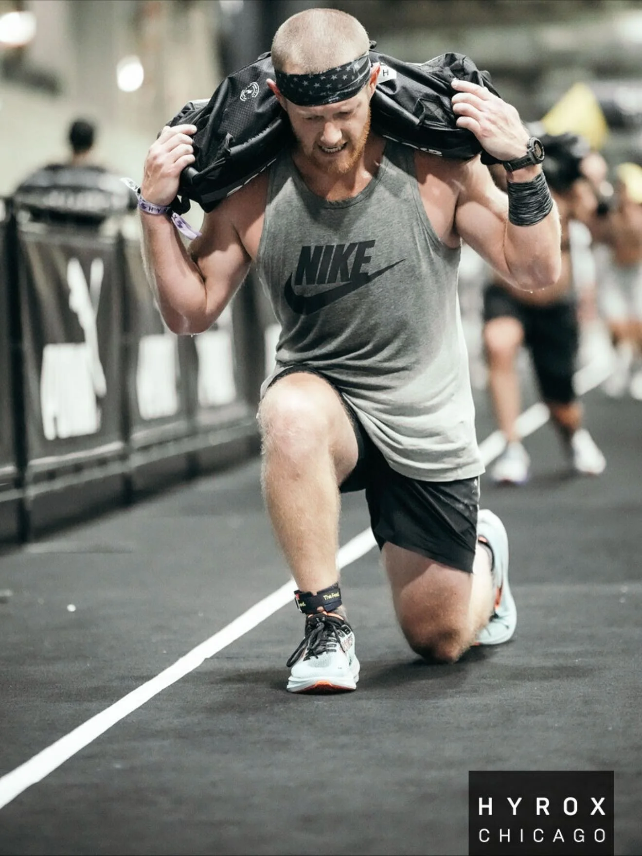 A muscular man wearing a gray Nike tank top, black shorts, and a bandana on his head is kneeling on one knee while carrying a black bag on his shoulders. The environment appears to be a gym or fitness center with other people in the background.