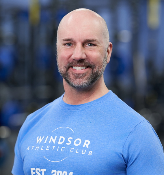 Man with a shaved head and beard smiling, wearing a blue Windsor Athletic Club t-shirt, standing in a gym with blurred equipment in the background.