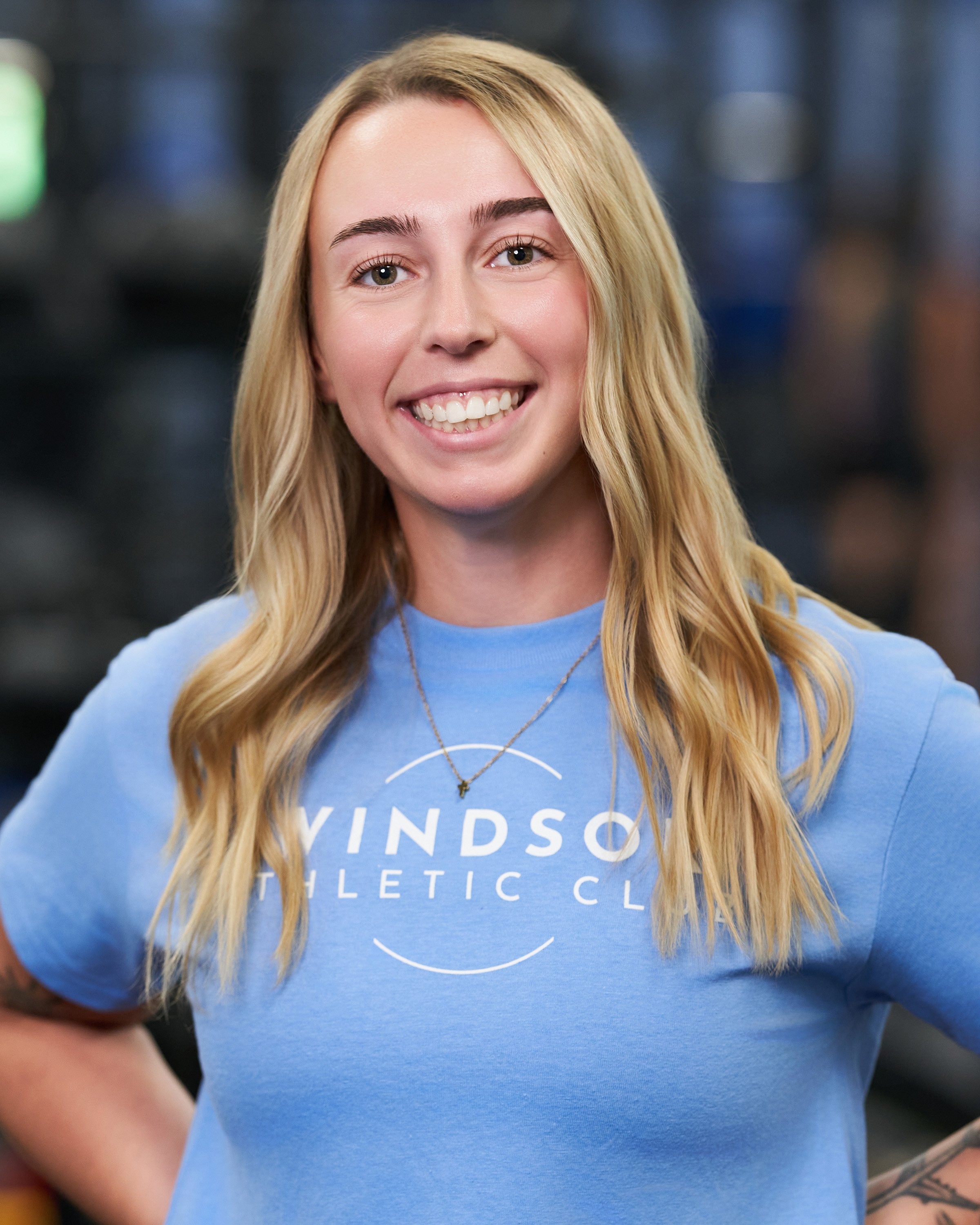 A young woman in a blue 'Windsor Athletic Club' T-shirt, smiling, with long dark hair, standing in a gym.