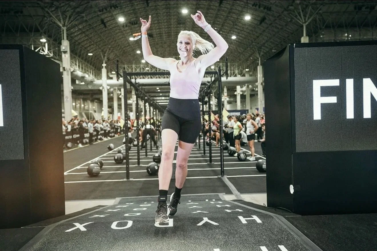 A woman celebrating crossing the finish line at a fitness competition inside a large gym with high ceilings, with kettlebells and people in the background.