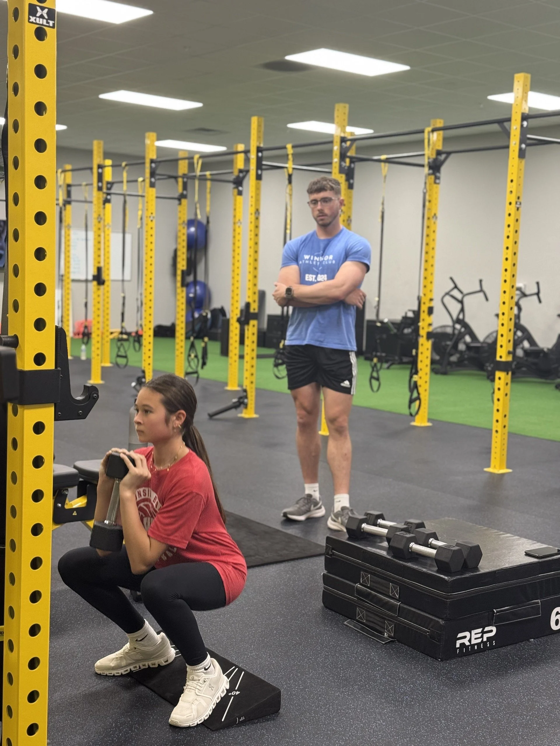 A young woman wearing a red t-shirt and black leggings is performing a squat exercise with a dumbbell across her shoulders, while an older man in athletic attire watches her in a gym with yellow and black workout equipment.
