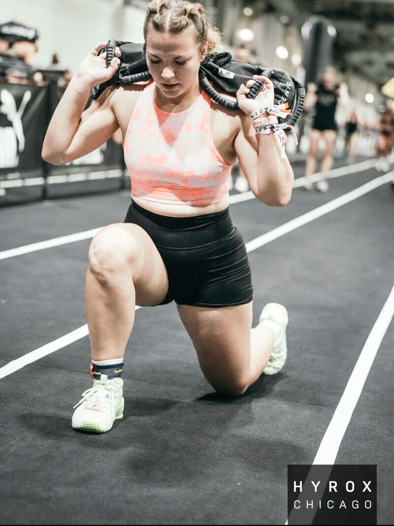 A woman in athletic wear kneels on one knee on a running track, pulling a weighted vest onto her shoulders during a workout at Hyrox Chicago.