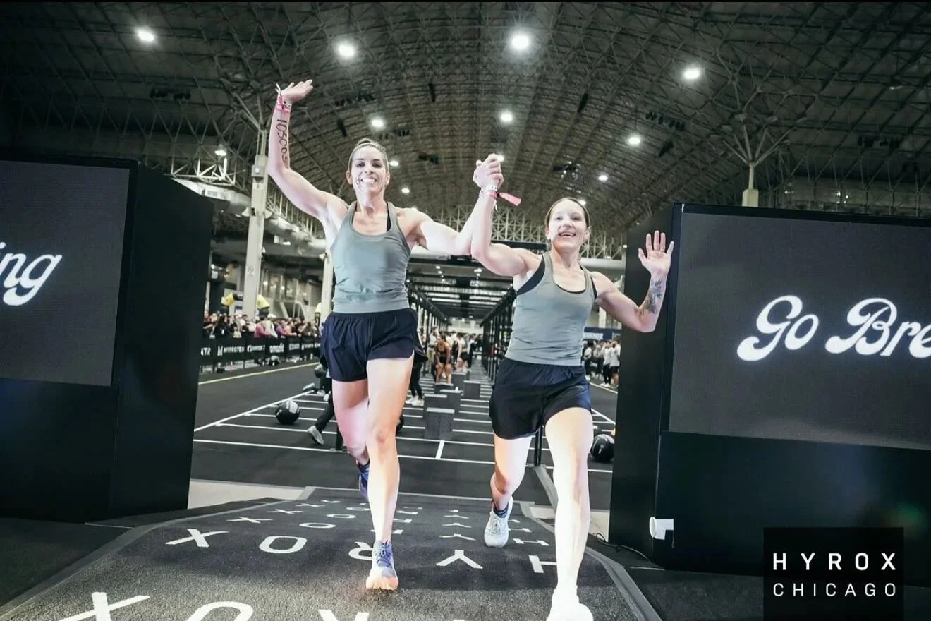 Two women in athletic clothing running through an indoor gym event, holding hands and smiling. Large signs with motivational messages are visible.