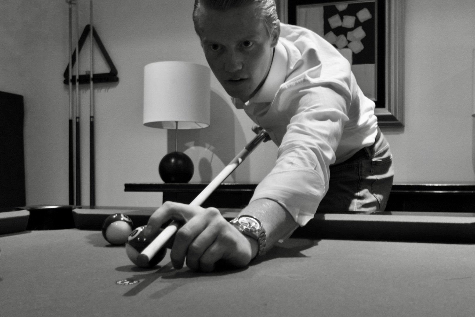 A man in a white shirt leaning over a pool table, aiming to hit the cue ball with a cue stick, in a room with a table lamp, wall art, and pool cues in the background. private mens style consultant boulder Colorado personal styling styled by swift