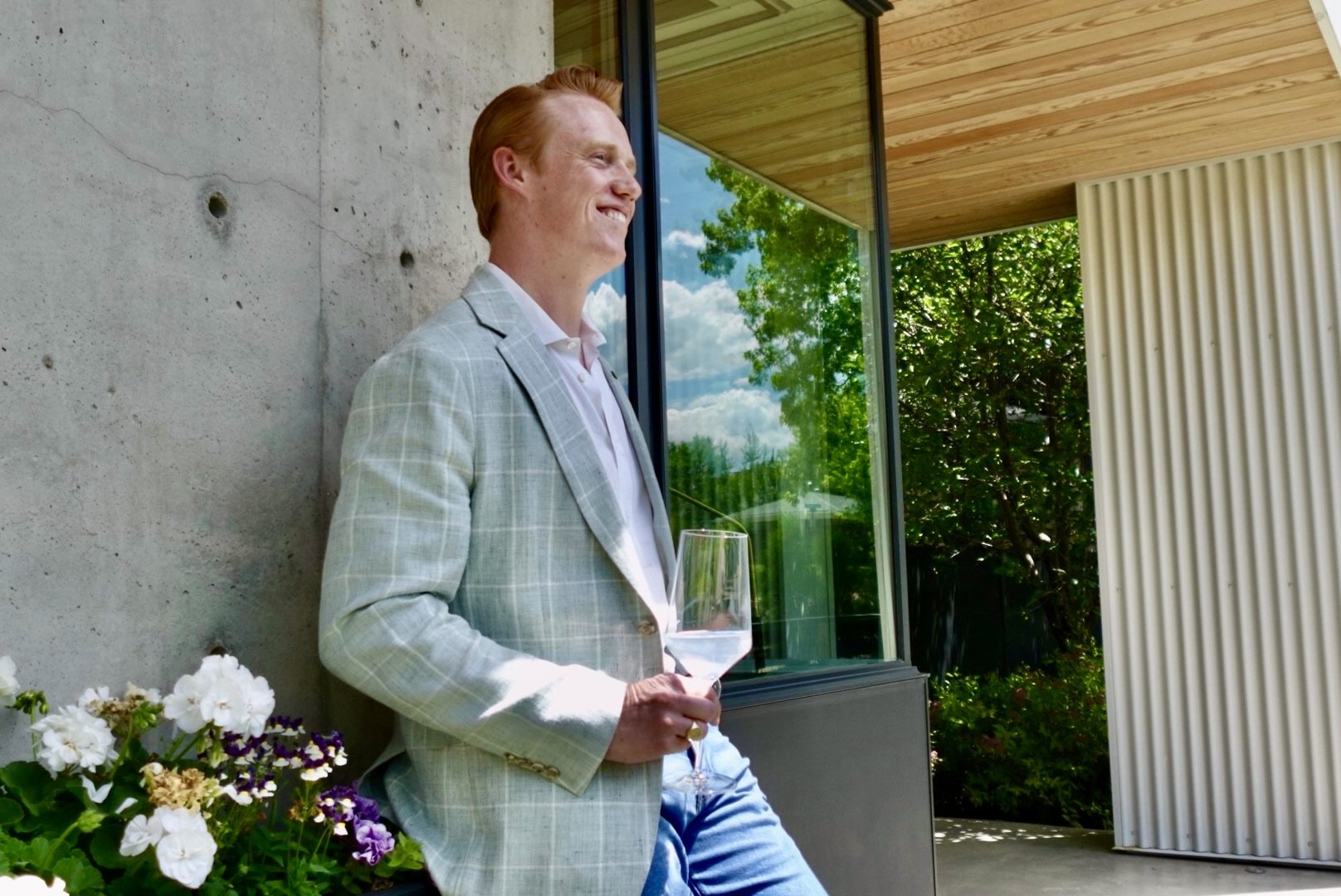 A man with red hair is standing outdoors, holding a glass of white wine, wearing a light gray checkered blazer and a white shirt, smiling and looking off to the side. private mens style consultant boulder Colorado personal styling styled by swift
