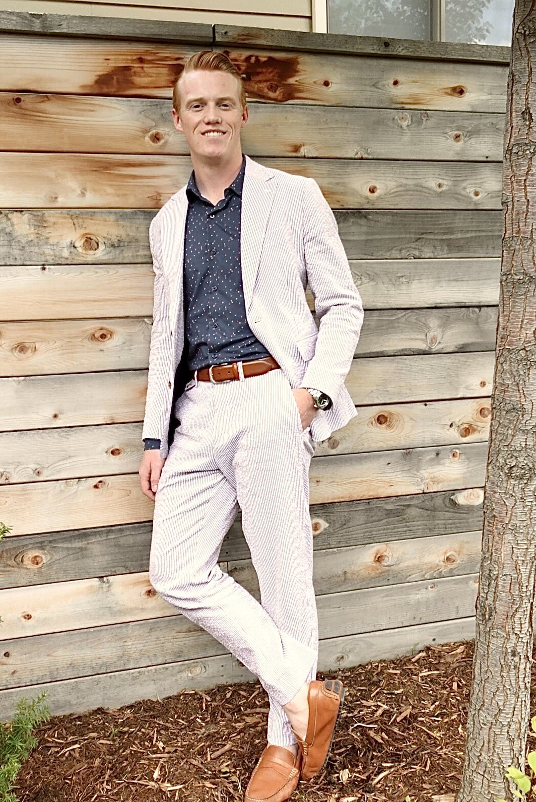 A man standing outdoors in front of a wooden fence, dressed in a seersucker pinstripe suit, navy shirt, brown shoes and belt, smiling hand in his pocket. private mens style consultant boulder Colorado personal styling styled by swift