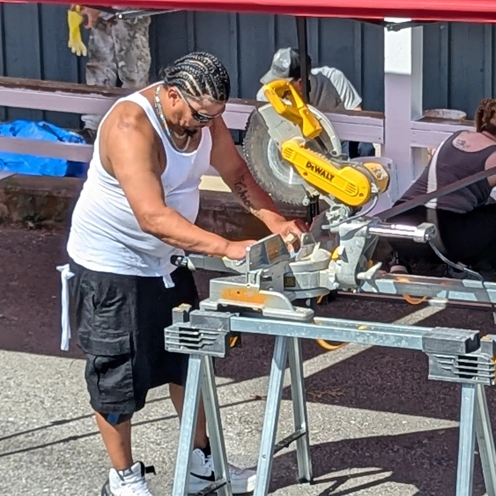 A man using a yellow DeWalt miter saw outdoors, standing at a work table, wearing a white tank top, black shorts, sunglasses, and a cap with braided hair.