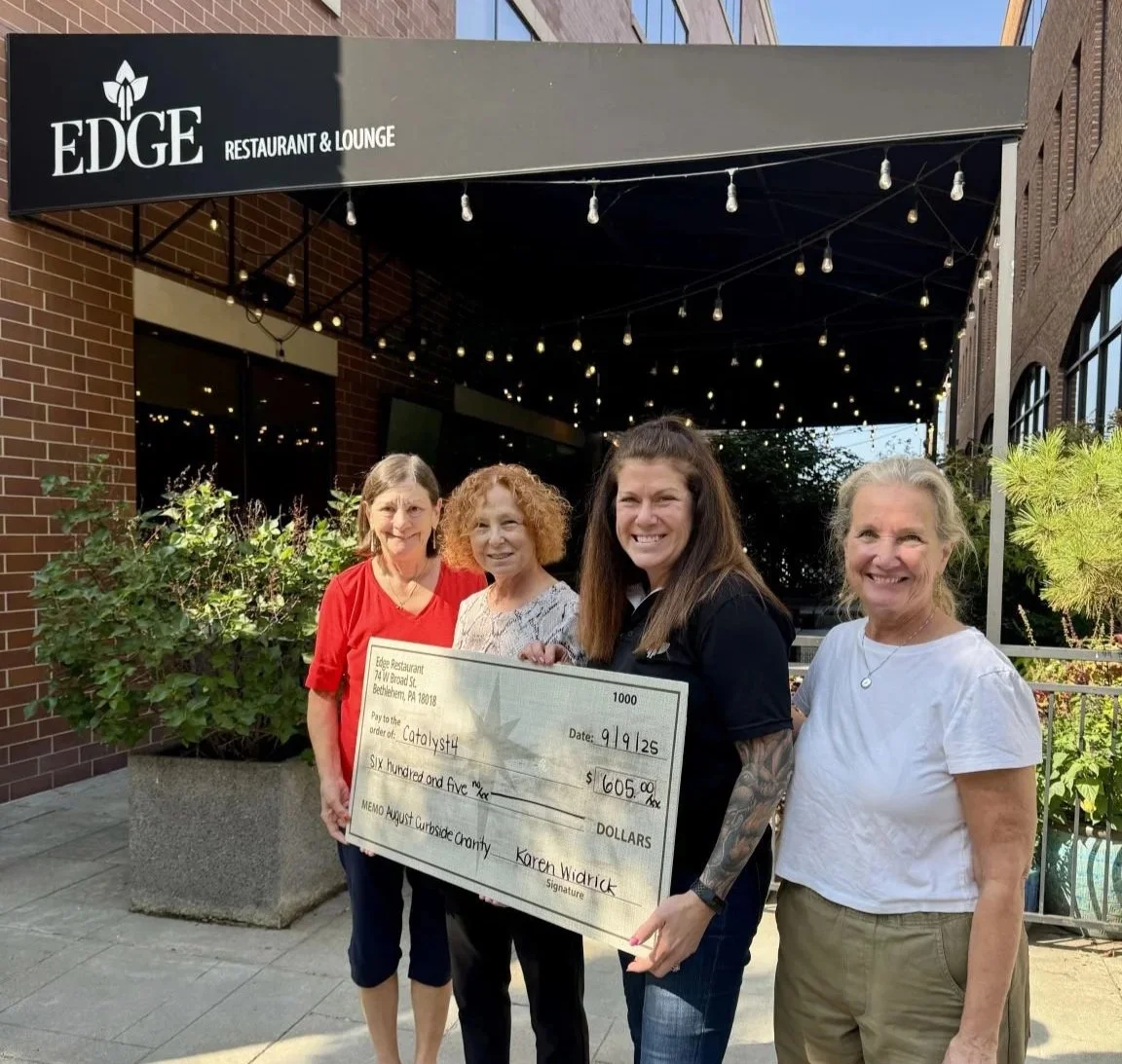 Four women standing outside a restaurant named Edge Restaurant & Lounge, holding a large check for $605 made out to Catalyst. The women are smiling, and there are string lights hanging overhead, with a brick building and green plants in the background.