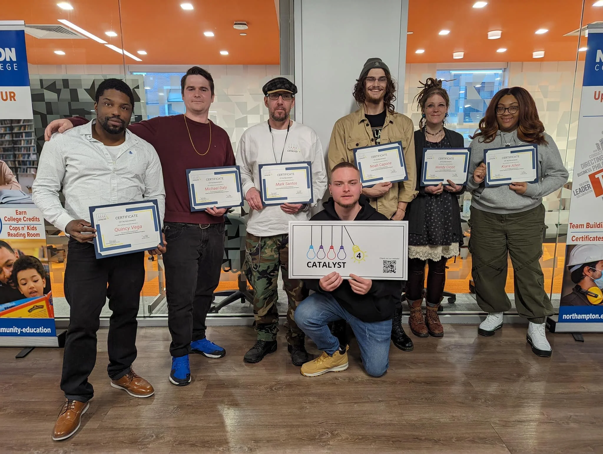 Group of seven people holding certificates in an indoor setting, with one person kneeling in front holding a sign that says 'CATALYST.'