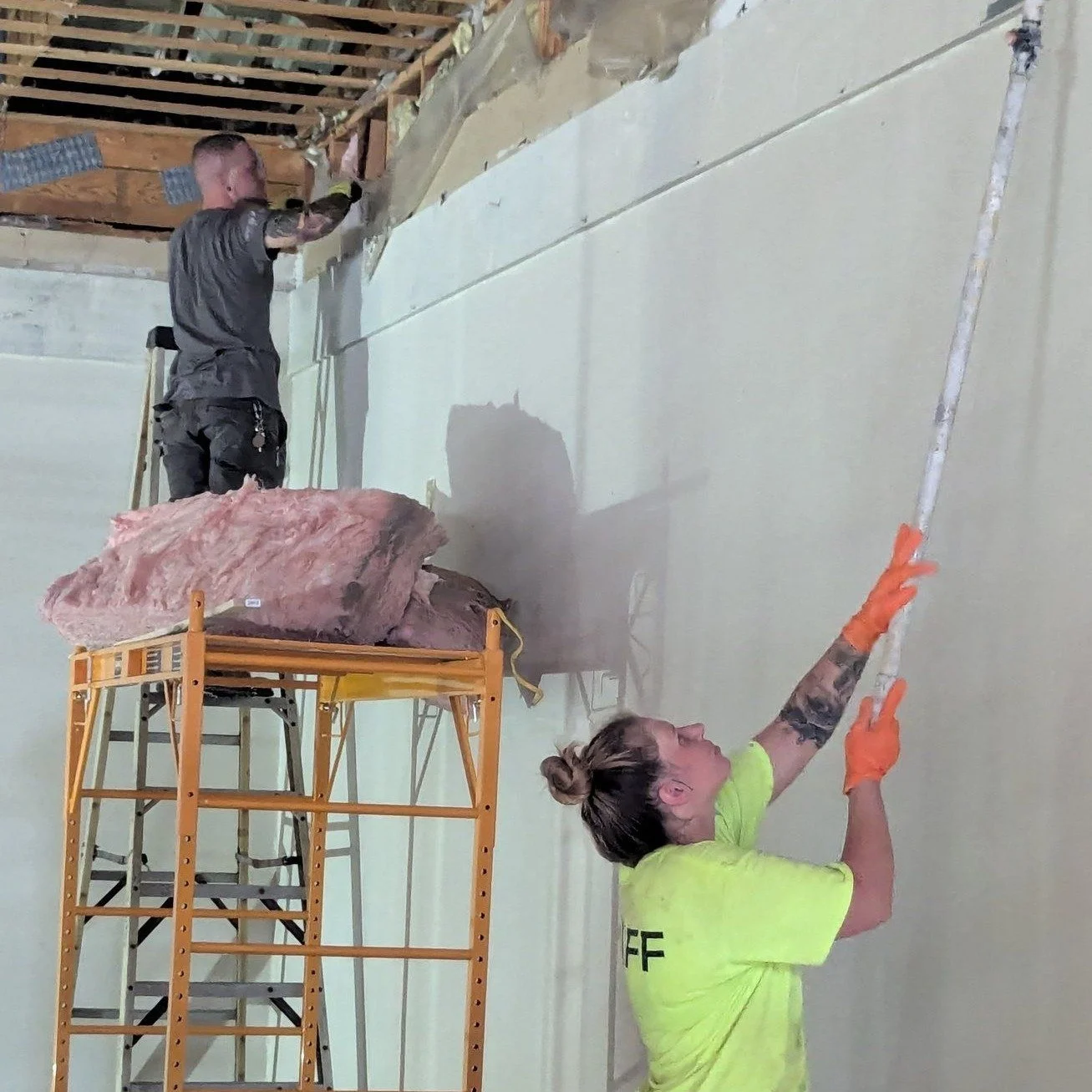 Two workers installing insulation on an interior wall of a building, with one woman reaching up with a tool and a man working higher on the wall.