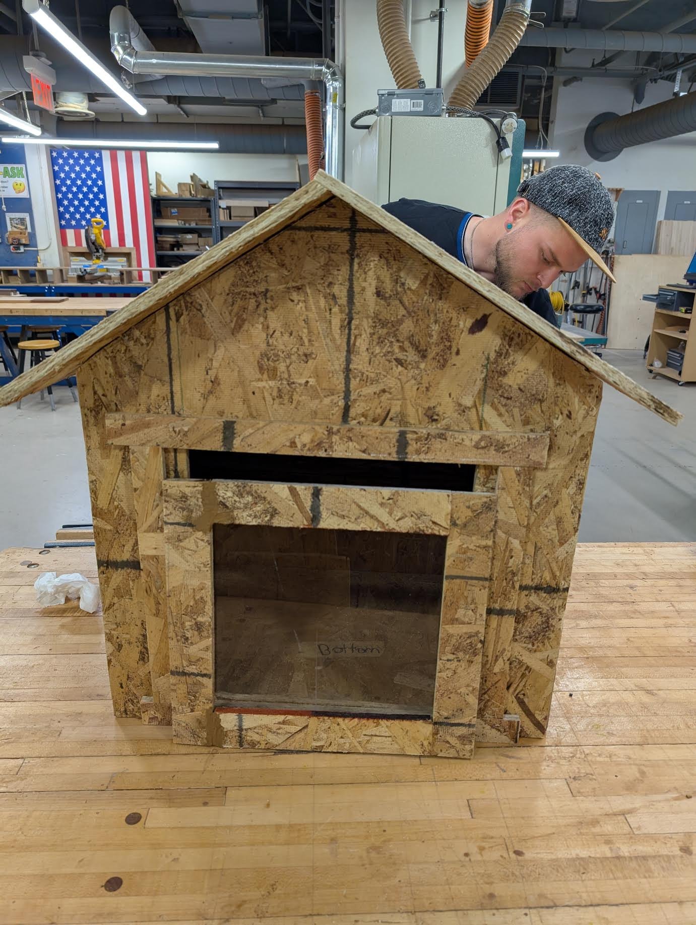 A person working on building a small wooden doghouse in a woodworking shop.