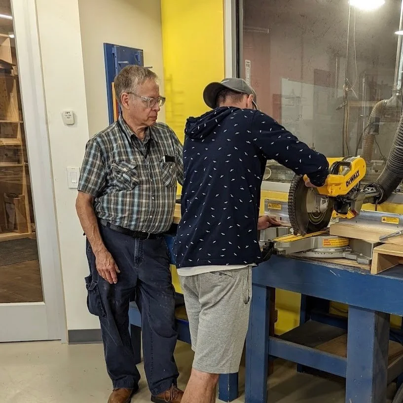 An older man observing a young person operating a yellow DeWalt cutting saw in a woodworking workshop.