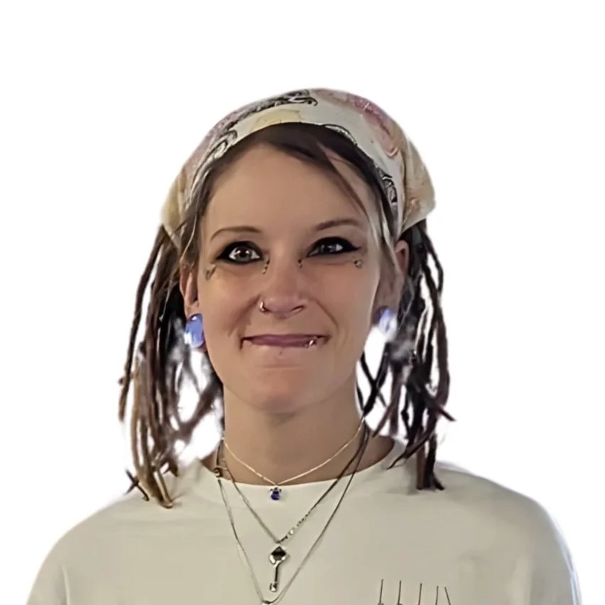 A woman with light skin and brown dreadlocks, wearing a colorful headscarf, multiple necklaces, earrings, and facial piercings, is making a playful facial expression.
