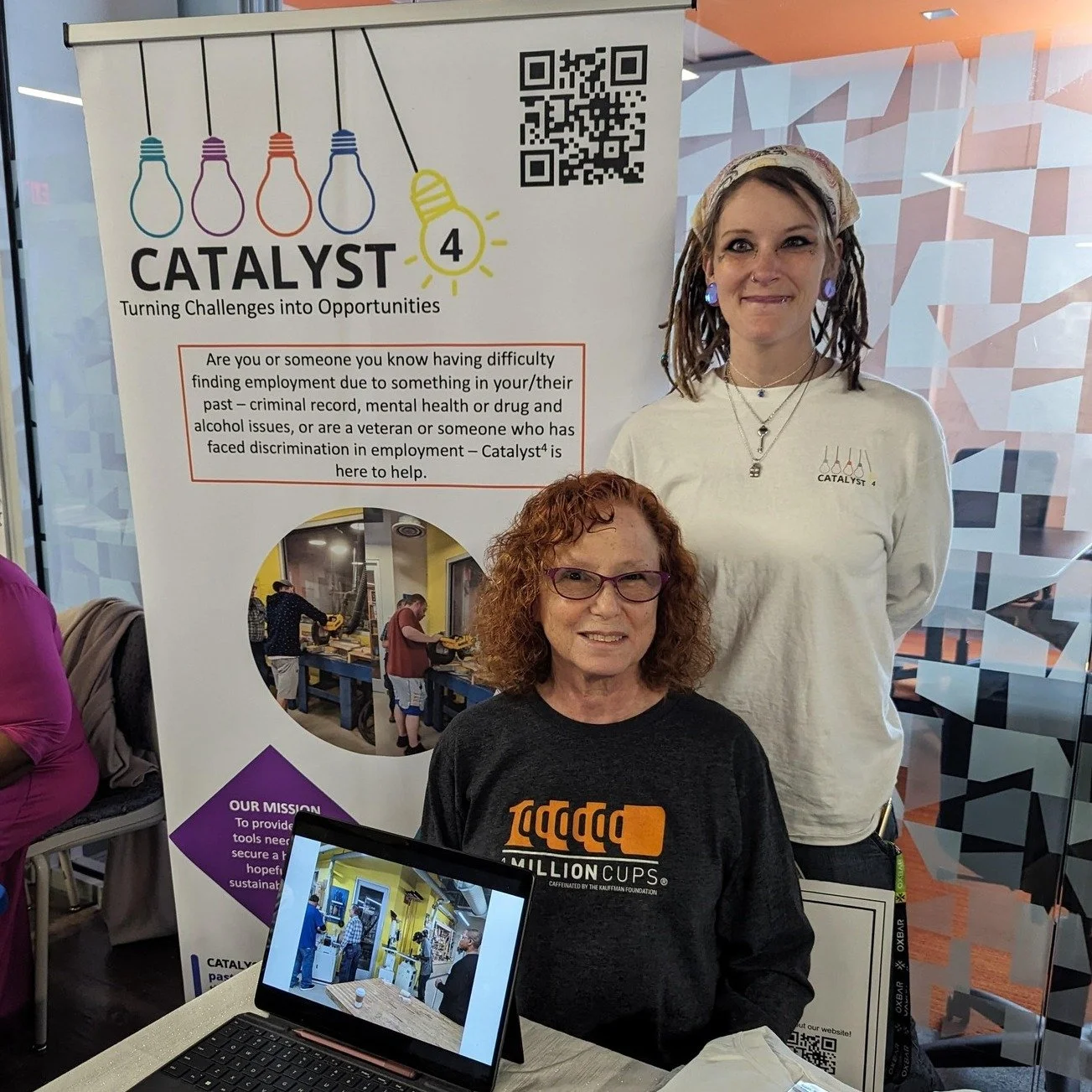 A woman with curly red hair and glasses sitting in front of a table with a laptop, at a booth with a banner that reads 'CATALYST 4: Turning Challenges into Opportunities.' Two women are standing behind her, one with dreadlocks and the other with a headscarf, smiling. The booth promotes assistance for employment challenges related to criminal records, mental health, substance issues, or veterans.