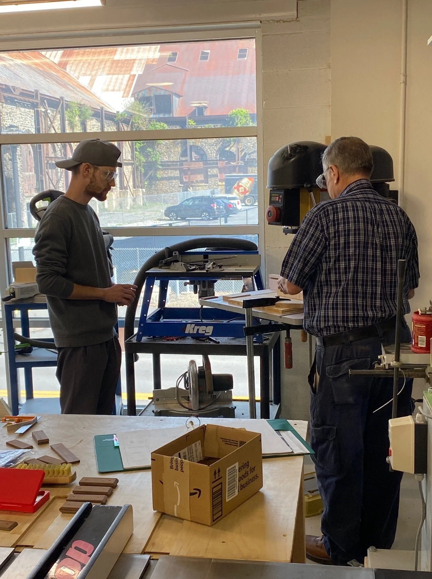 Two men working in a woodworking shop, one operating a machine and the other standing nearby, with woodworking tools and papers on a table in the foreground.