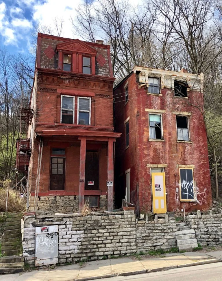 An old, abandoned multi-story brick building with boarded-up windows, graffiti, and deteriorating exterior, situated on a street with a brick retaining wall and stairs.