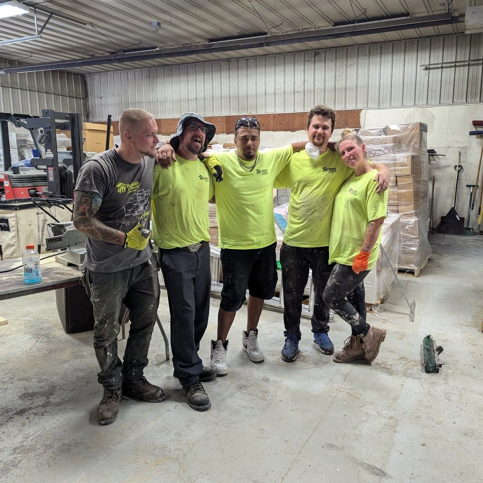 Group of five construction workers standing together in a warehouse, with tools and boxes in the background, smiling and posing for the photo.
