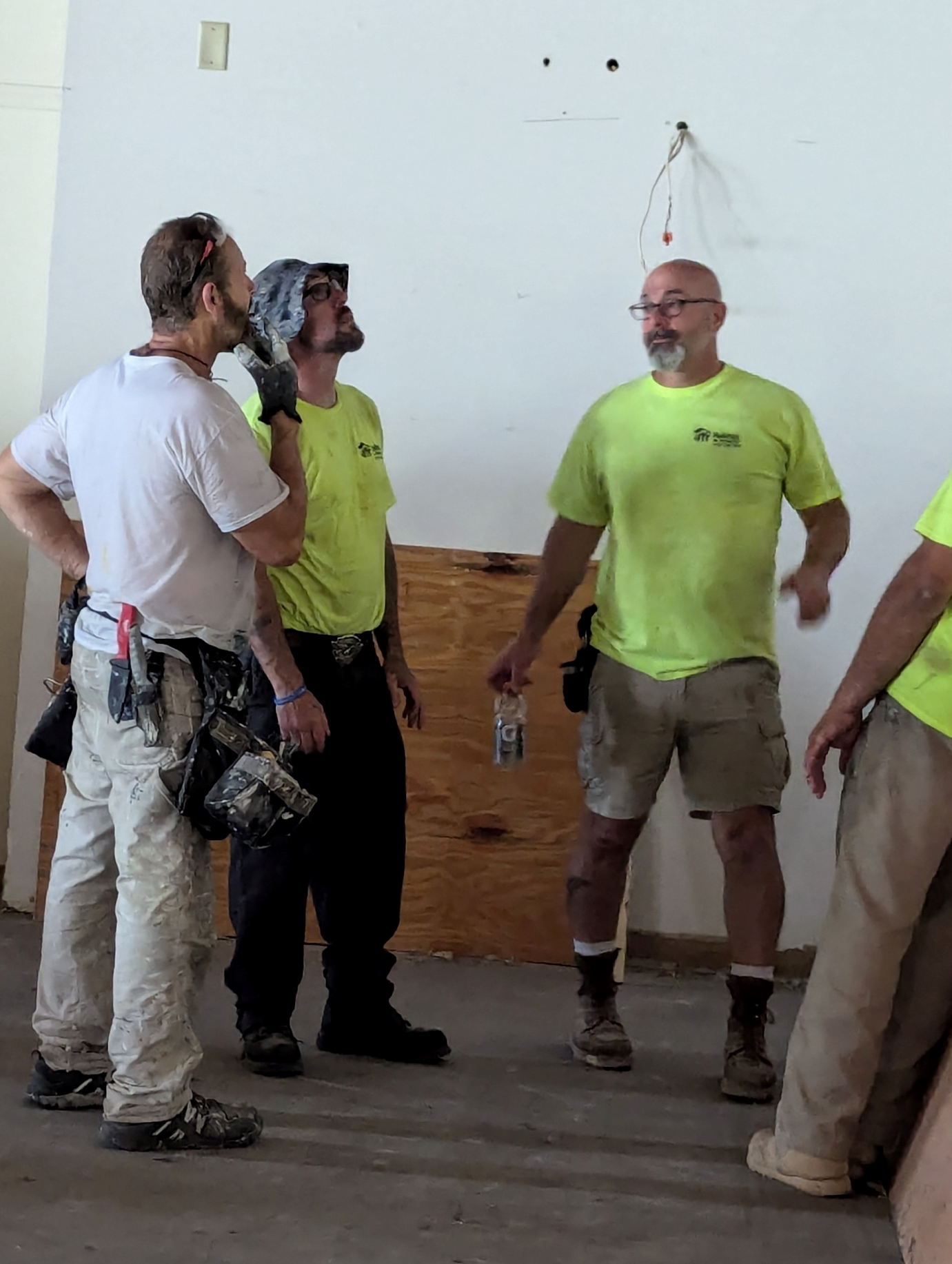 Group of construction workers in a room during a break, some with tools and wearing work gear, standing near a blank wall and a wooden panel.