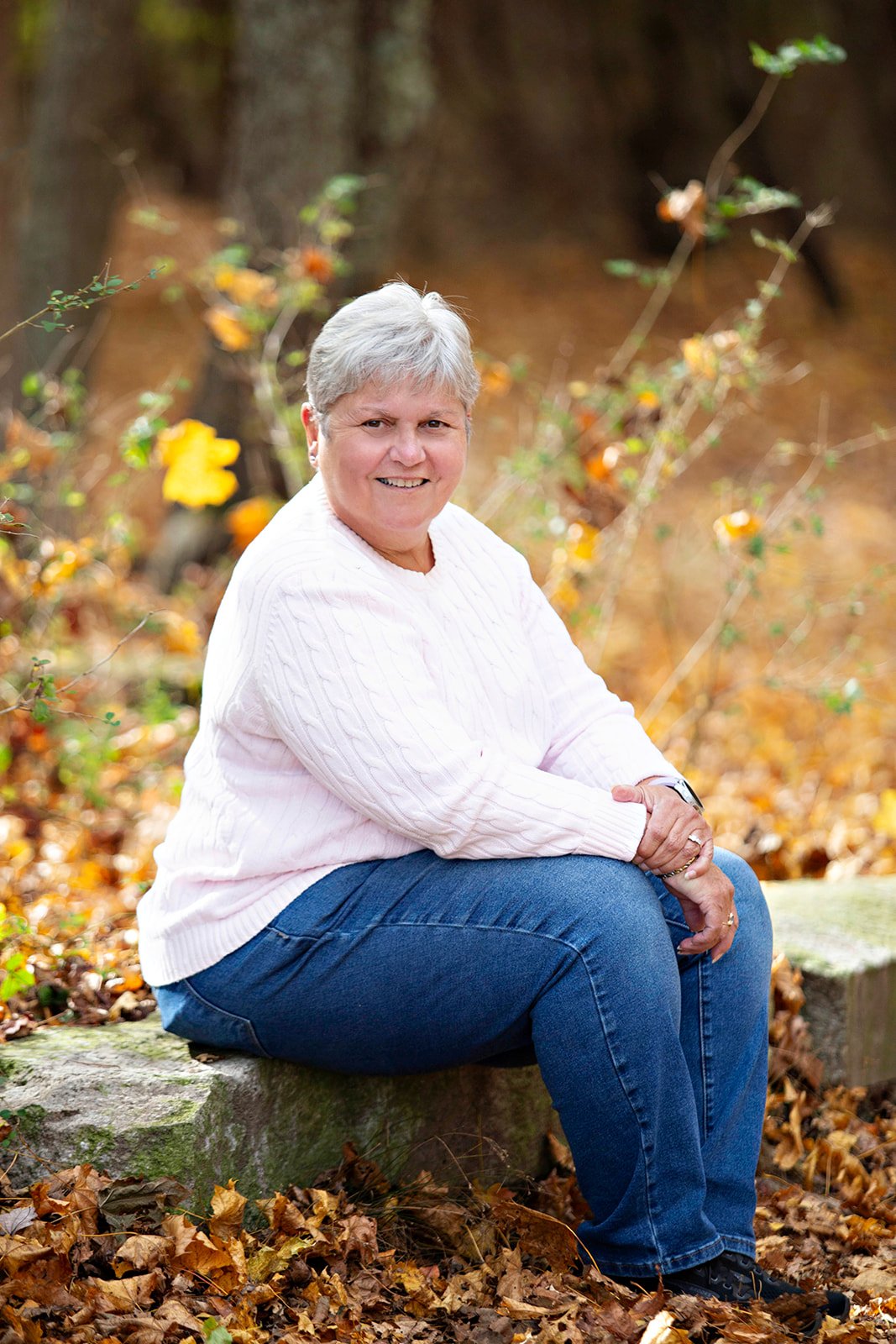 Joanne Cook sitting on a rock in a forest during autumn, surrounded by fallen leaves and trees.