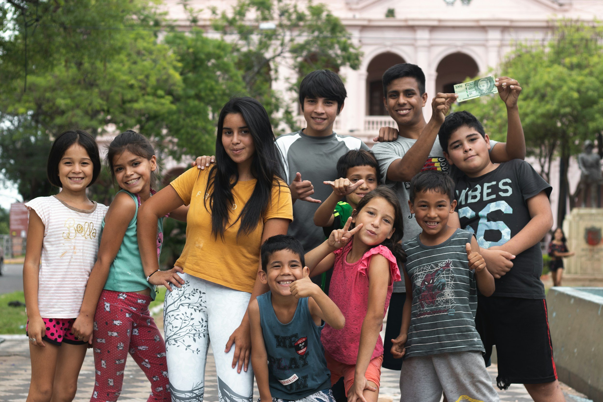 Group of smiling children and teenagers outdoors, some making peace signs, with a large building and trees in the background.