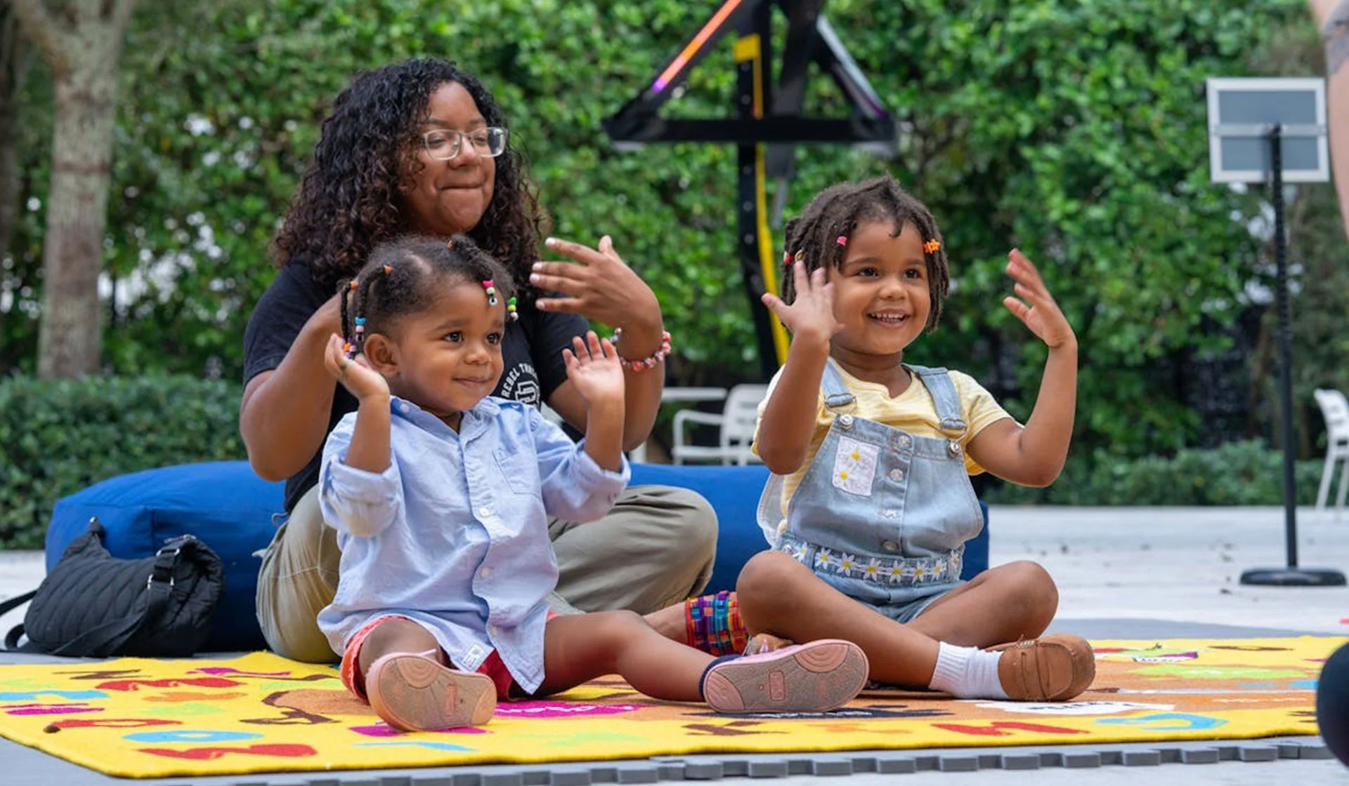 A woman with glasses and curly hair sitting on a colorful mat outdoors, playing with two smiling young girls who have dreadlocks and are wearing casual clothes. The girls are raising their hands and appear to be engaged in a fun activity.