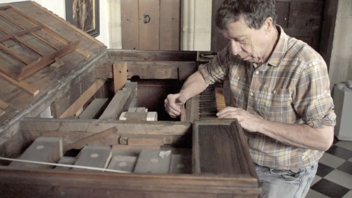 A man in a plaid shirt working on the inside of an old upright piano in a rustic room.