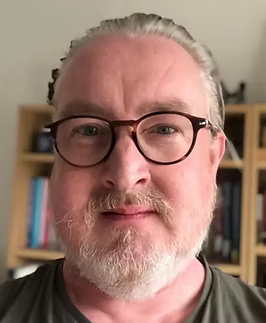 A close-up of a middle-aged man with gray hair, glasses, and a beard, standing in front of a bookshelf.