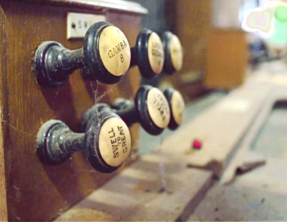 Close-up of vintage brass and black switches and knobs labeled with names, mounted on a wooden panel, with cobwebs and dust, in an old, possibly abandoned, mechanical or control room.