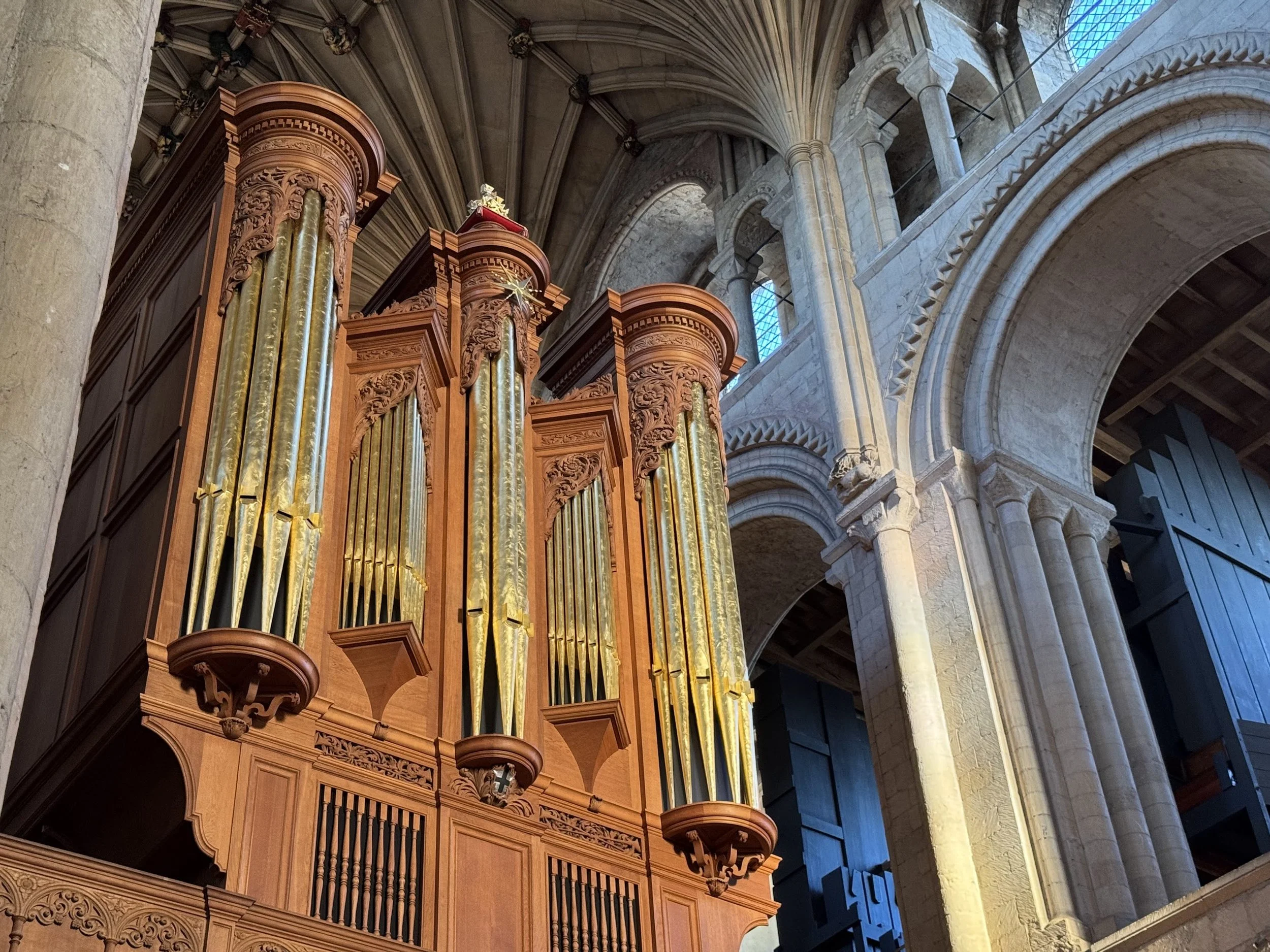 A large wooden pipe organ with golden pipes inside a stone cathedral with high vaulted ceilings and ornate stone arches.
