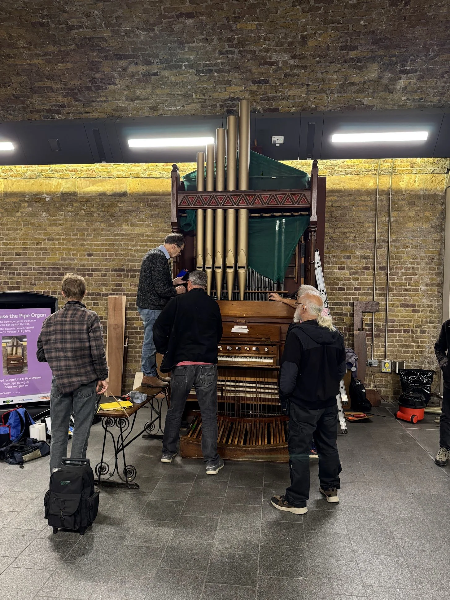 A group of people gathered around an old pipe organ in an indoor setting with a brick wall background.