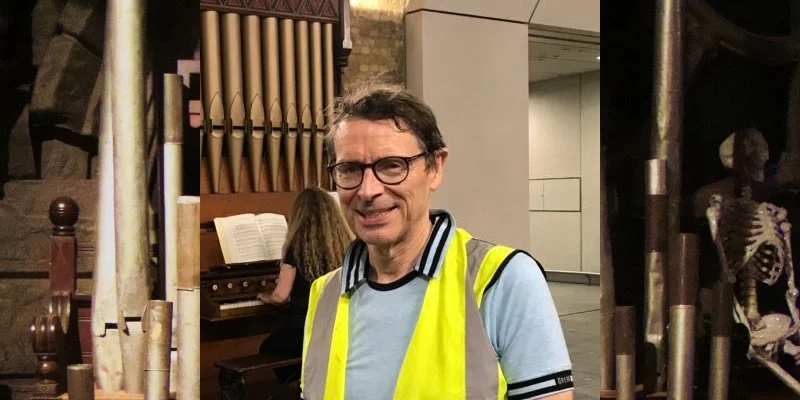 A man in a yellow safety vest standing indoors near a woman playing an organ, with a skeleton in the background.