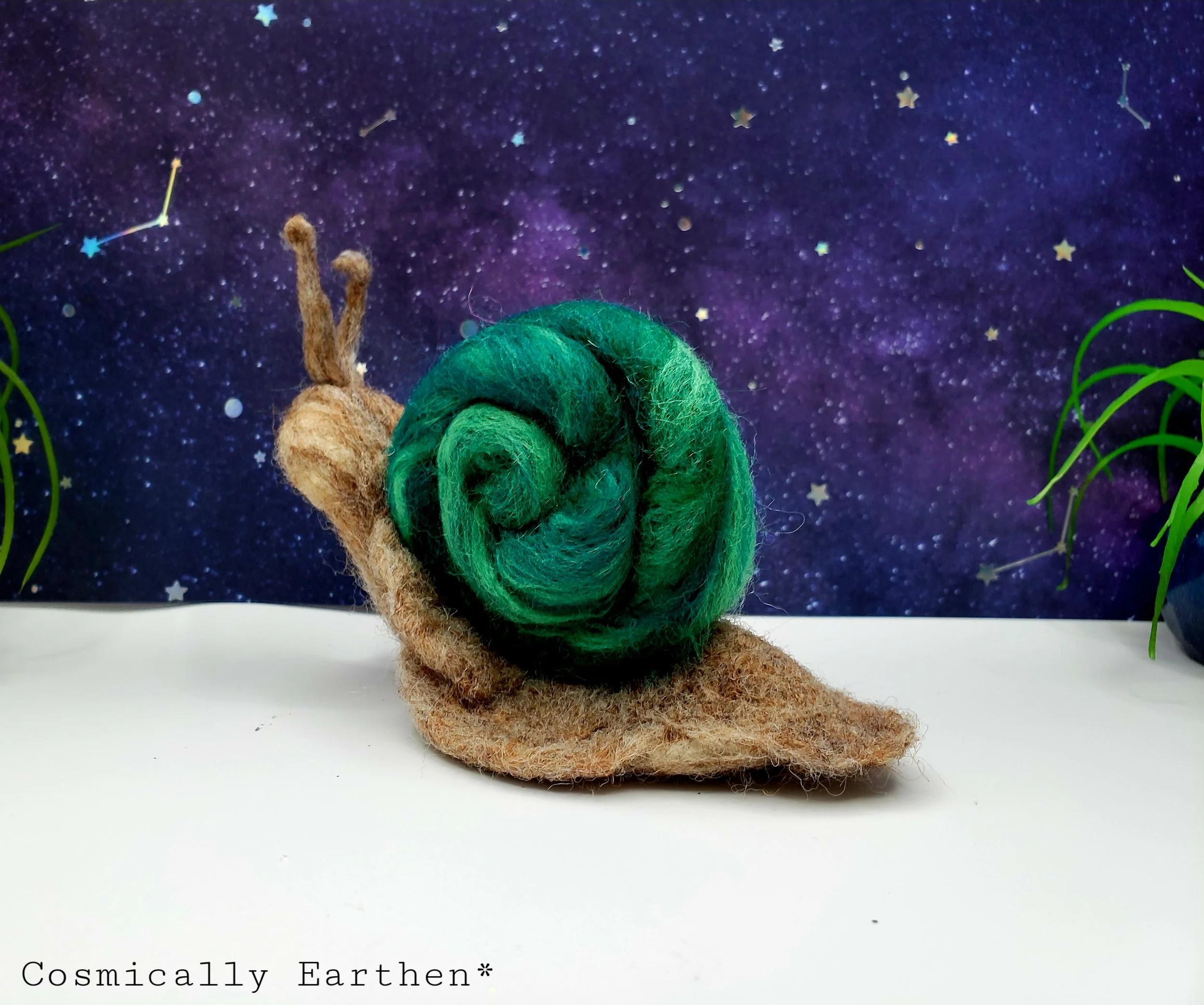 A felt snail with a green shell and a brown body, set against a starry space background with some plants on either side.