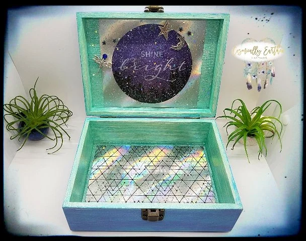 Open light blue wooden box with glittery mirrored interior, surrounded by potted green plants and wall decor with celestial and inspirational themes.